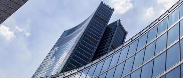 Modern office building with a curved glass façade and a high-rise tower seen from below.