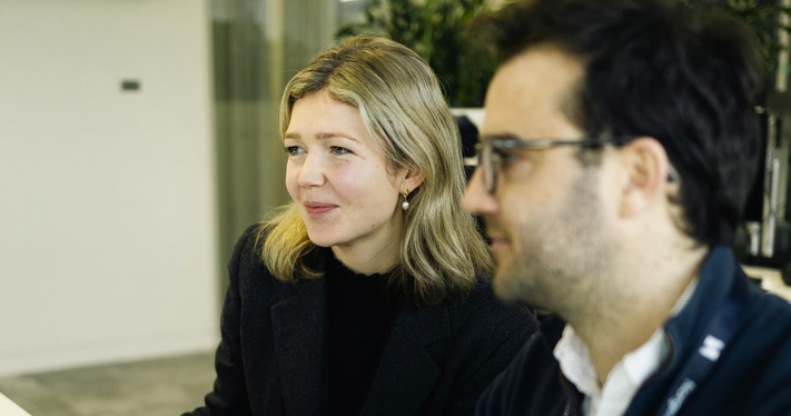 Two employees in conversation at a desk in an office.